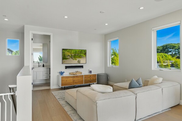 A modern living room with a large sectional sofa, a flat-screen TV mounted on the wall, and a view of greenery through the windows.