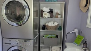 A compact laundry room with two stacked front-loading washing machines, a drying rack, a shelf with folded towels, a basket, and a small sink with a mirror.