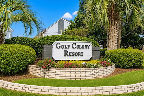 A sign for Golf Colony Resort surrounded by well-maintained landscaping and a clear blue sky.