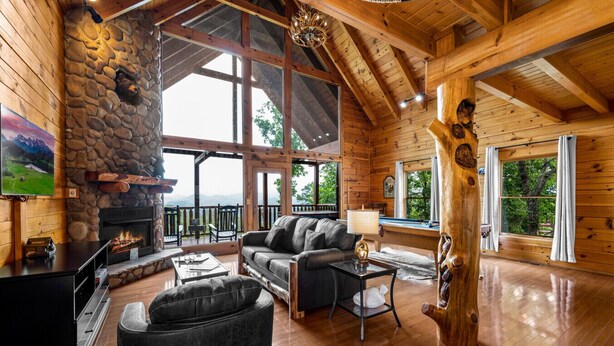 A cozy log cabin interior with a stone fireplace, leather furniture, and large windows offering scenic views. - Romantic Lookout | Incredible Views - Hot Tub - Pool Table (Sevierville)