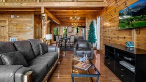 A cozy log cabin interior with a leather sofa, a dining table, and a TV mounted on the wall. - Romantic Lookout | Incredible Views - Hot Tub - Pool Table (Sevierville)