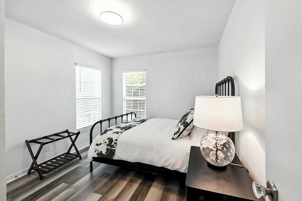 A bedroom with a bed, a bedside table with a lamp, a bench, and a window with blinds.