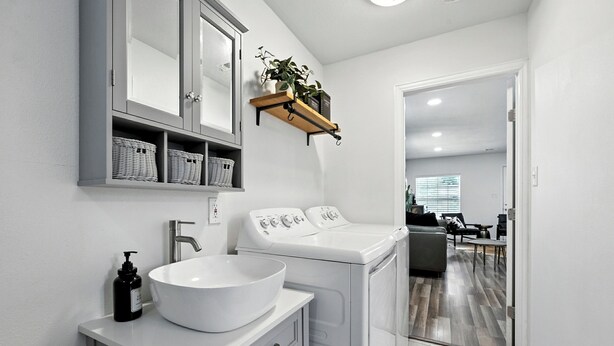 A modern bathroom with a white sink, a mirror, and a washing machine. There is a shelf with baskets and a plant above the sink. - Boots & Bedrolls - Walkable to Fort Worth's Dickies Arena. (Fort Worth)
