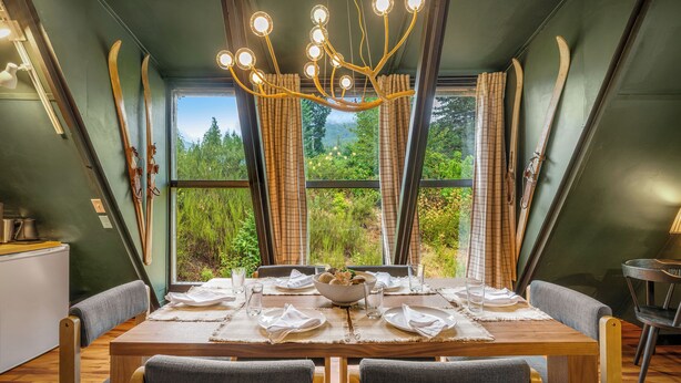 A dining area with a wooden table set for four, large windows with curtains, and skis hanging on the wall. - Packwood: Chalet + Nest (Packwood)