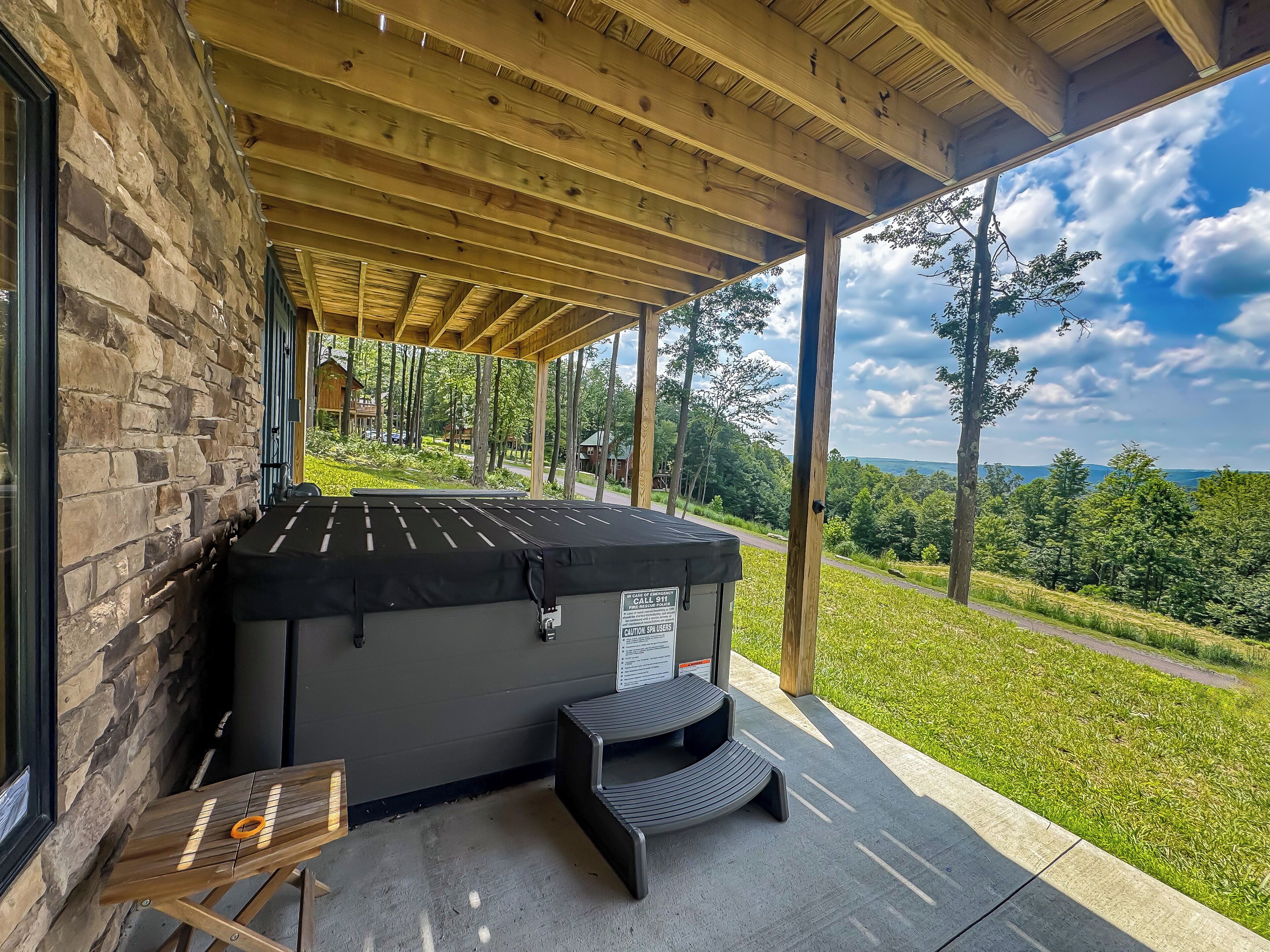 A hot tub under a wooden pergola with a stone wall backdrop, a small table, and a view of a green landscape.