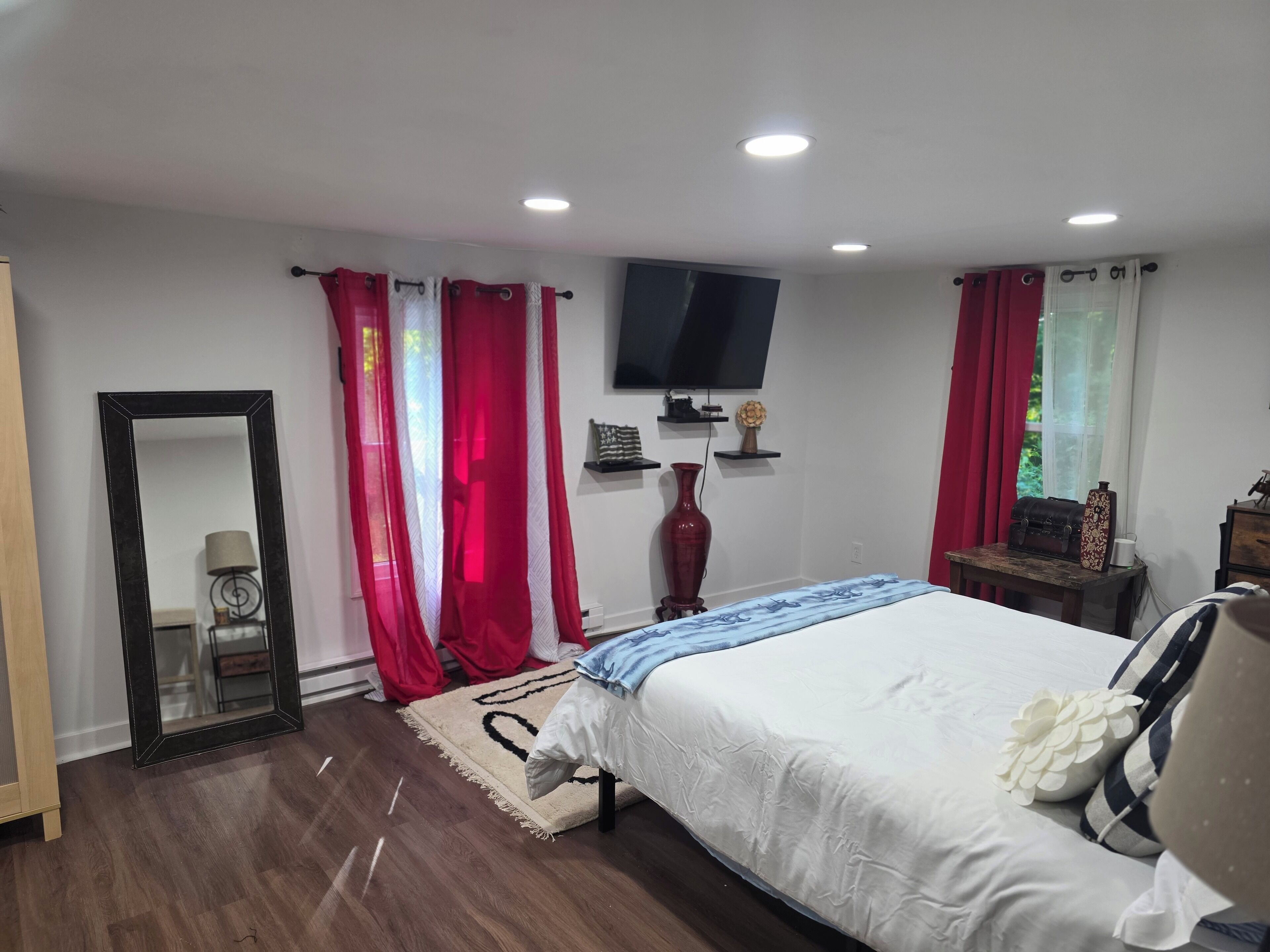 Een slaapkamer met een bed, een aan de muur bevestigde televisie, een spiegel en een raam met rode gordijnen.