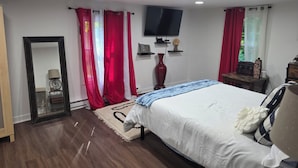 A bedroom with a bed, a TV mounted on the wall, a mirror, and a window with red curtains.