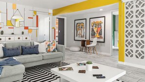 A modern living room with a grey sofa, a white coffee table, and a black and white striped rug. There are two framed artworks on the wall, a white cabinet, and a yellow accent wall.
