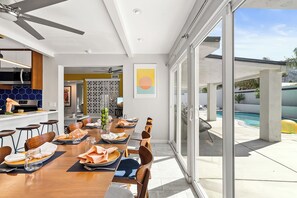Un espace repas moderne avec une longue table dressée pour six personnes, une cuisine équipée d’un four à micro-ondes et une vue sur une piscine.