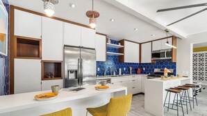 A modern kitchen with white cabinets, a stainless steel refrigerator, a built-in oven, and a blue tiled backsplash. There are two yellow chairs and a bar area with stools.