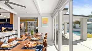 A modern dining area with a long table set for six, a kitchen with a microwave, and a view of a swimming pool.