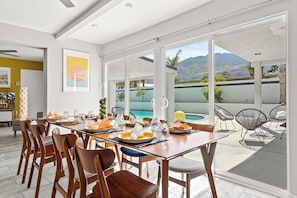 Uma sala de jantar com mesa posta para quatro pessoas, grandes janelas e vista para uma piscina e montanhas.
