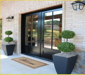 Une entrée d’immeuble avec une porte noire, deux plantes ornementales en pot et un paillasson.