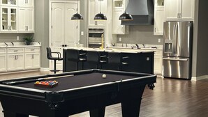 Eine moderne Küche mit weißen Schränken, einem Edelstahlkühlschrank, einem Billardtisch und Pendelleuchten.