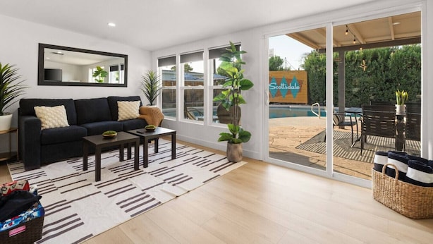 A modern living room with a sofa, coffee table, and potted plants. There is a large sliding glass door leading to an outdoor patio with a pool and seating area.