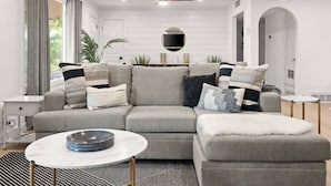A modern living room with a grey sectional sofa, a round coffee table, and a white armchair. There is a small side table with a bowl on it, and a patterned rug on the floor.