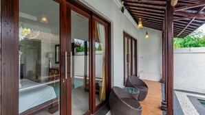 A room with a bed, a glass door leading to a pool, wicker chairs, and a ceiling with hanging lights. - Sakya Villa (Canggu)