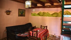 Une pièce avec un canapé en cuir noir, une table à manger en bois avec des chaises, un luminaire suspendu, un cactus fixé au mur et une fresque représentant des montagnes verdoyantes.
