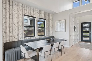 Un comedor moderno con una larga mesa, asientos tipo banca acolchados y ventanas grandes con vistas al exterior.