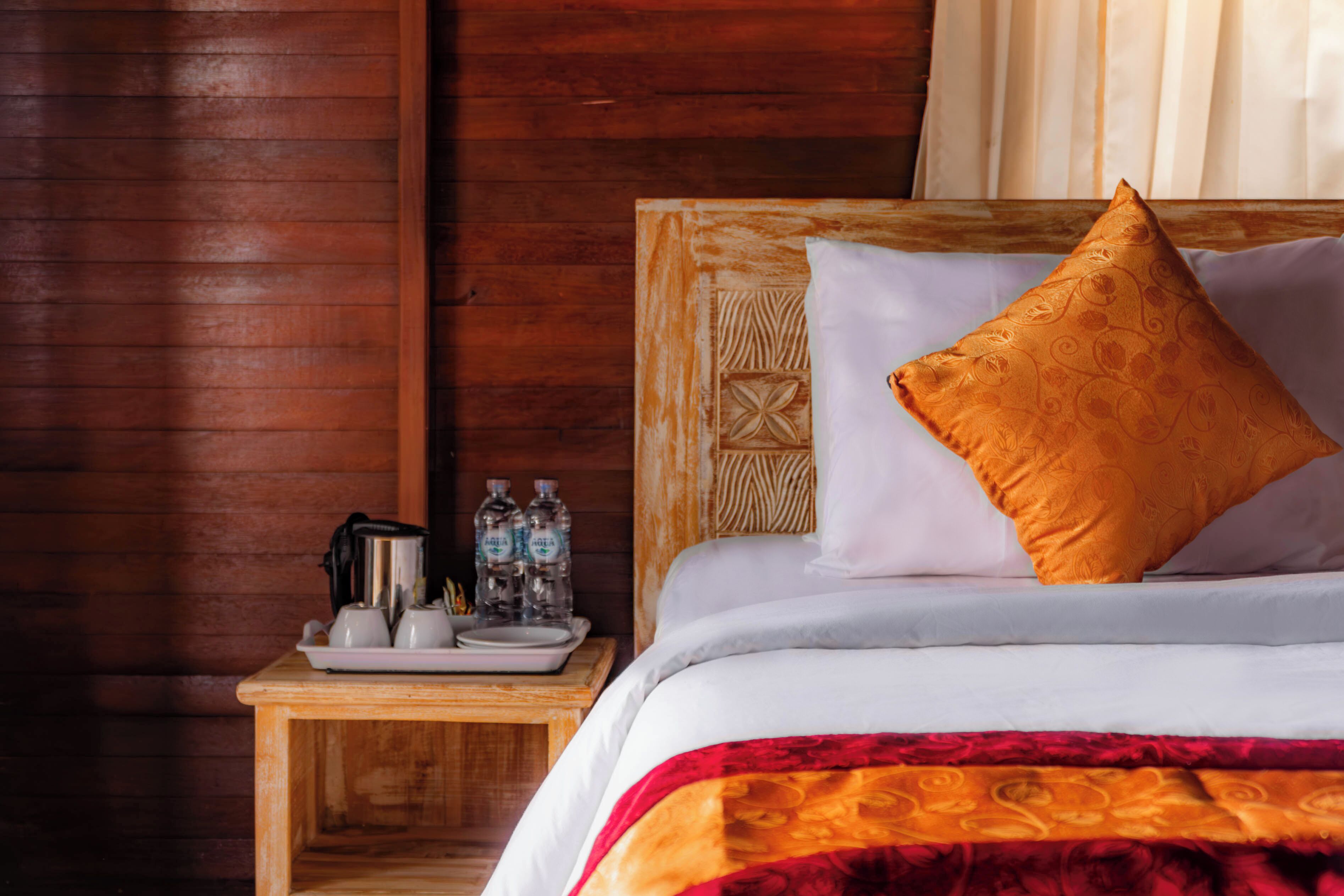 Una cama de madera con almohadas blancas y anaranjadas, una mesita de noche con un neceser, y una pared de madera.