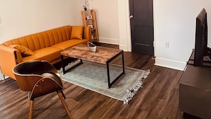 Een woonkamer met een bank, salontafel en een fauteuil. Er is een deur en een televisie in de kamer.