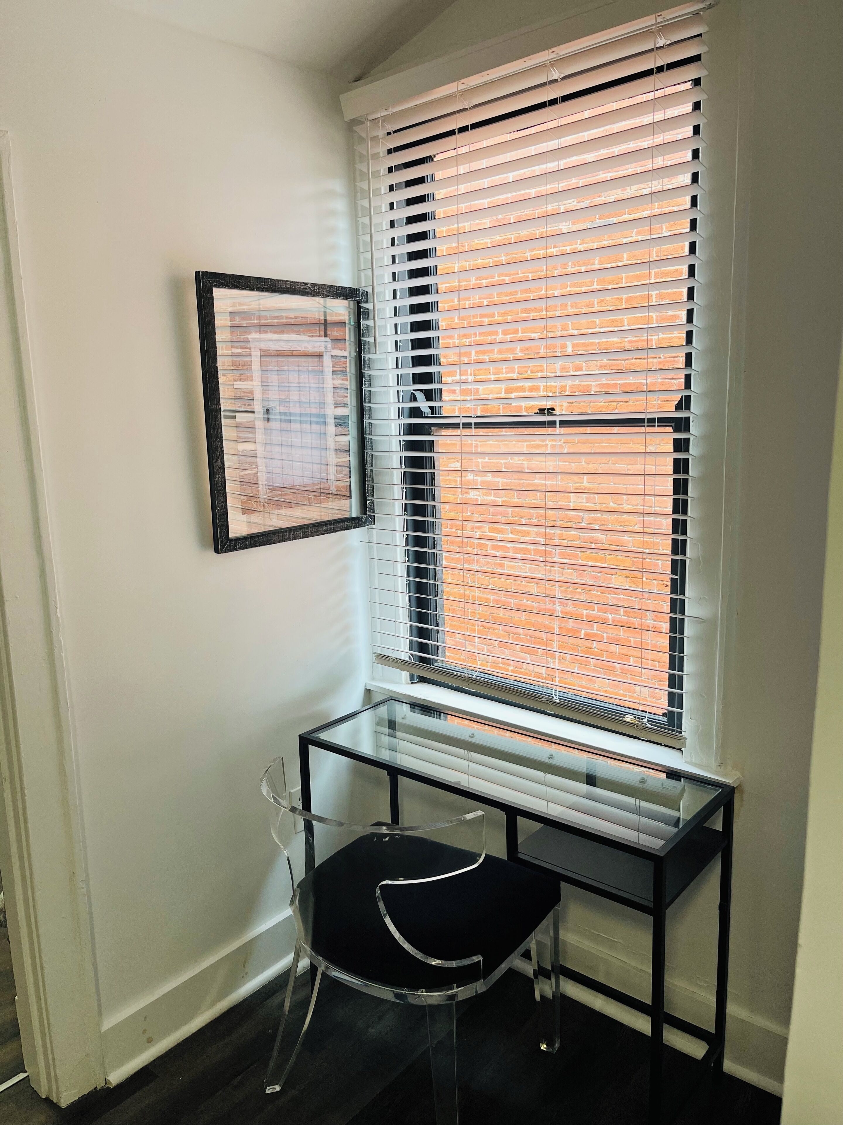 A corner room with a window, a framed picture on the wall, and a glass table with a chair.
