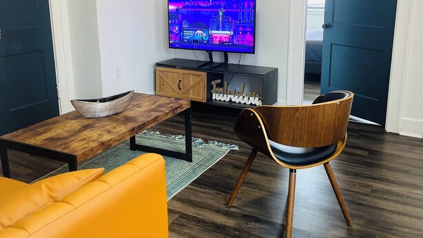 Een moderne woonkamer met een flatscreen televisie, een houten salontafel, een gele bank en een houten stoel.