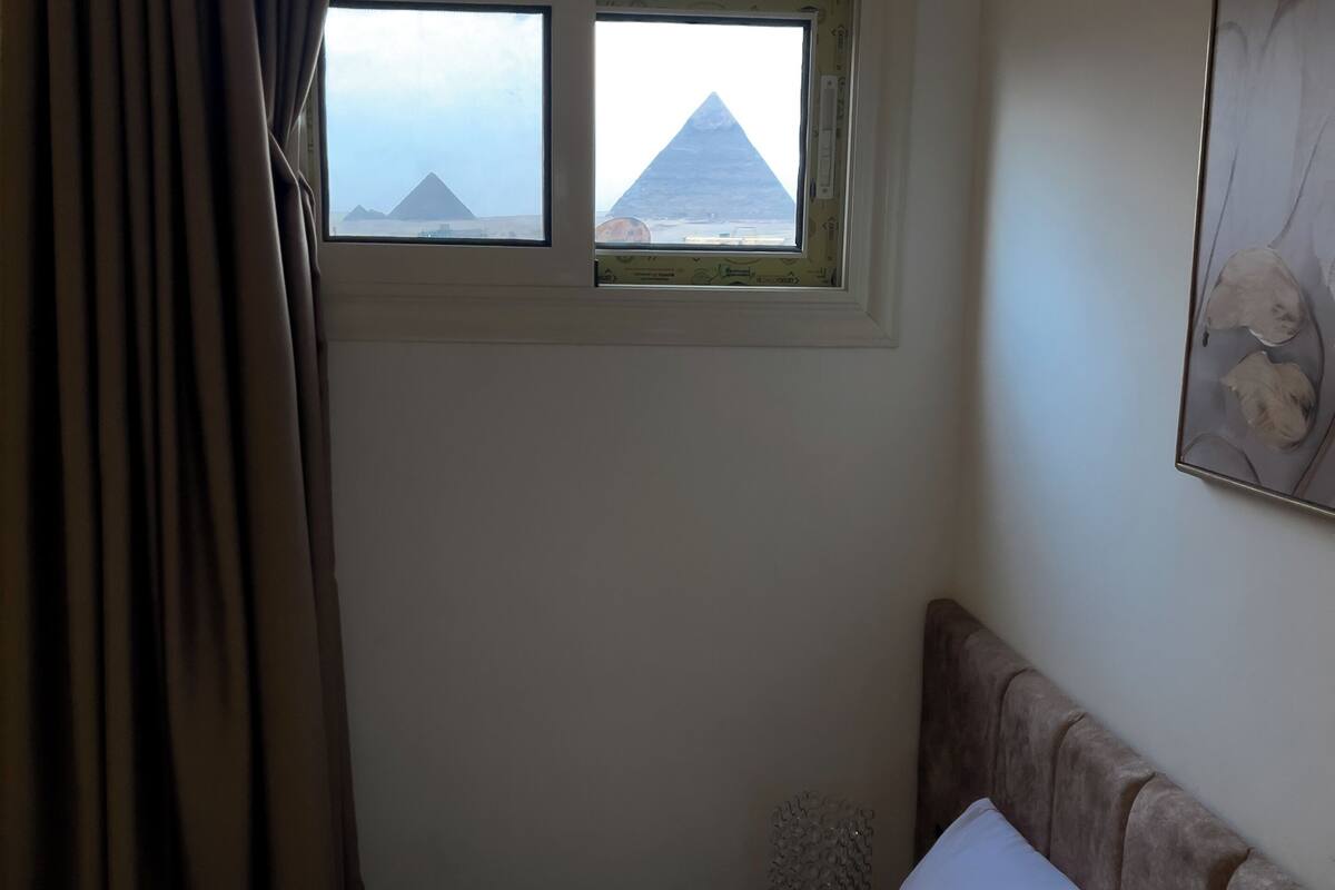 Ett hotellrum med en säng, ett nattduksbord, ett fönster med utsikt över pyramiderna och en gardin.