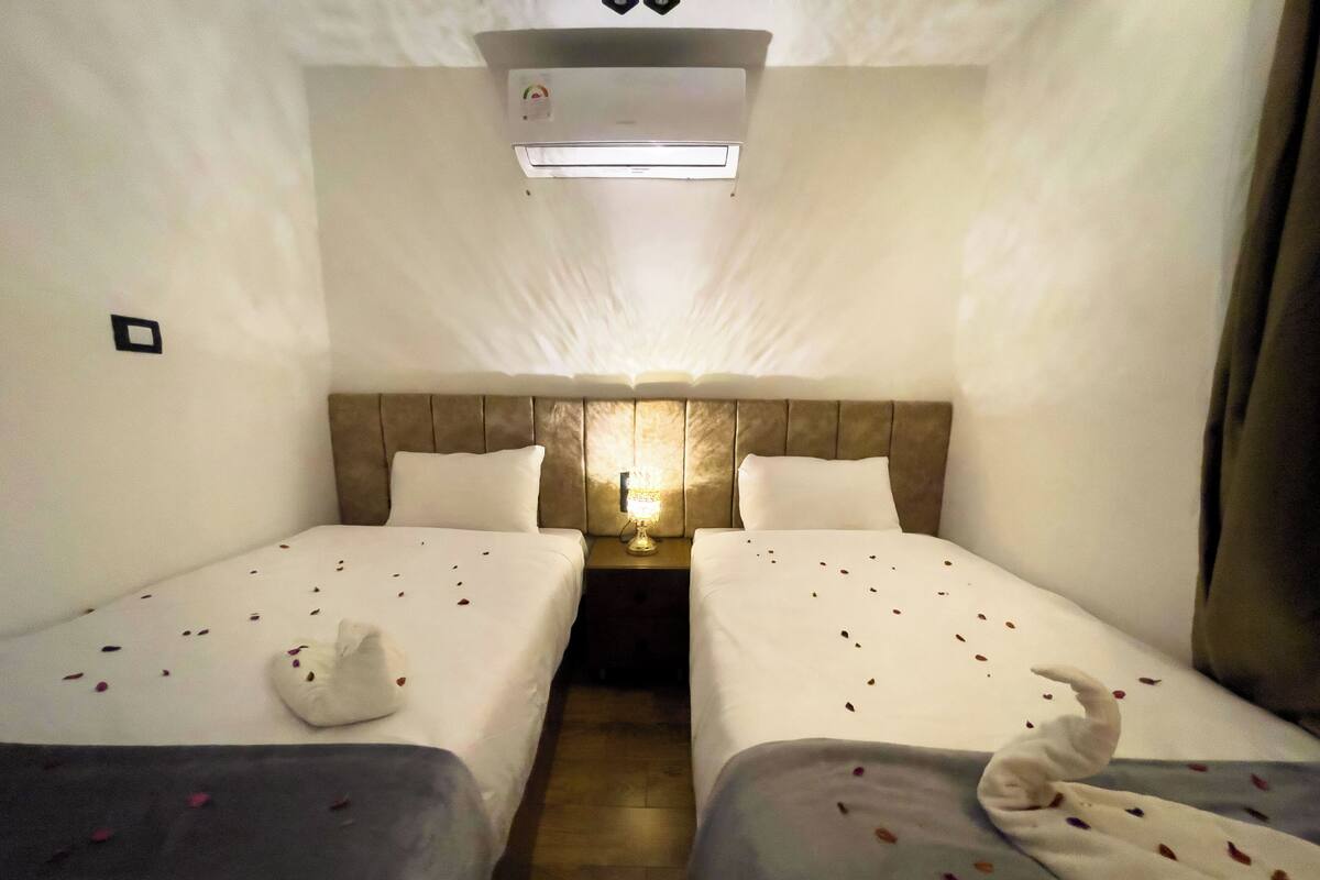 Ett hotellrum med två sängar, ett nattduksbord med en lampa, en luftkonditioneringsanläggning och ett litet fönster.