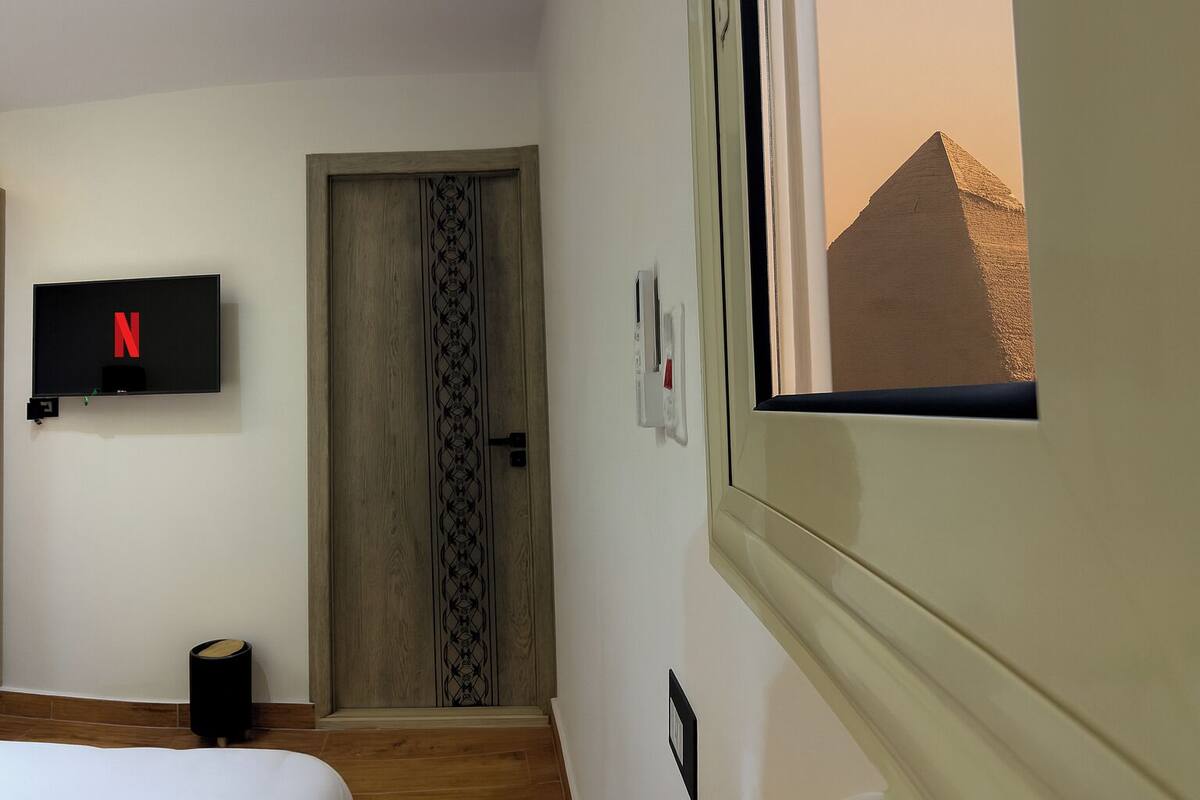 Ett hotellrum med en tv som är monterad på väggen, en trädörr med en dekorativ panel, en liten papperskorg och ett fönster med utsikt över en pyramid.