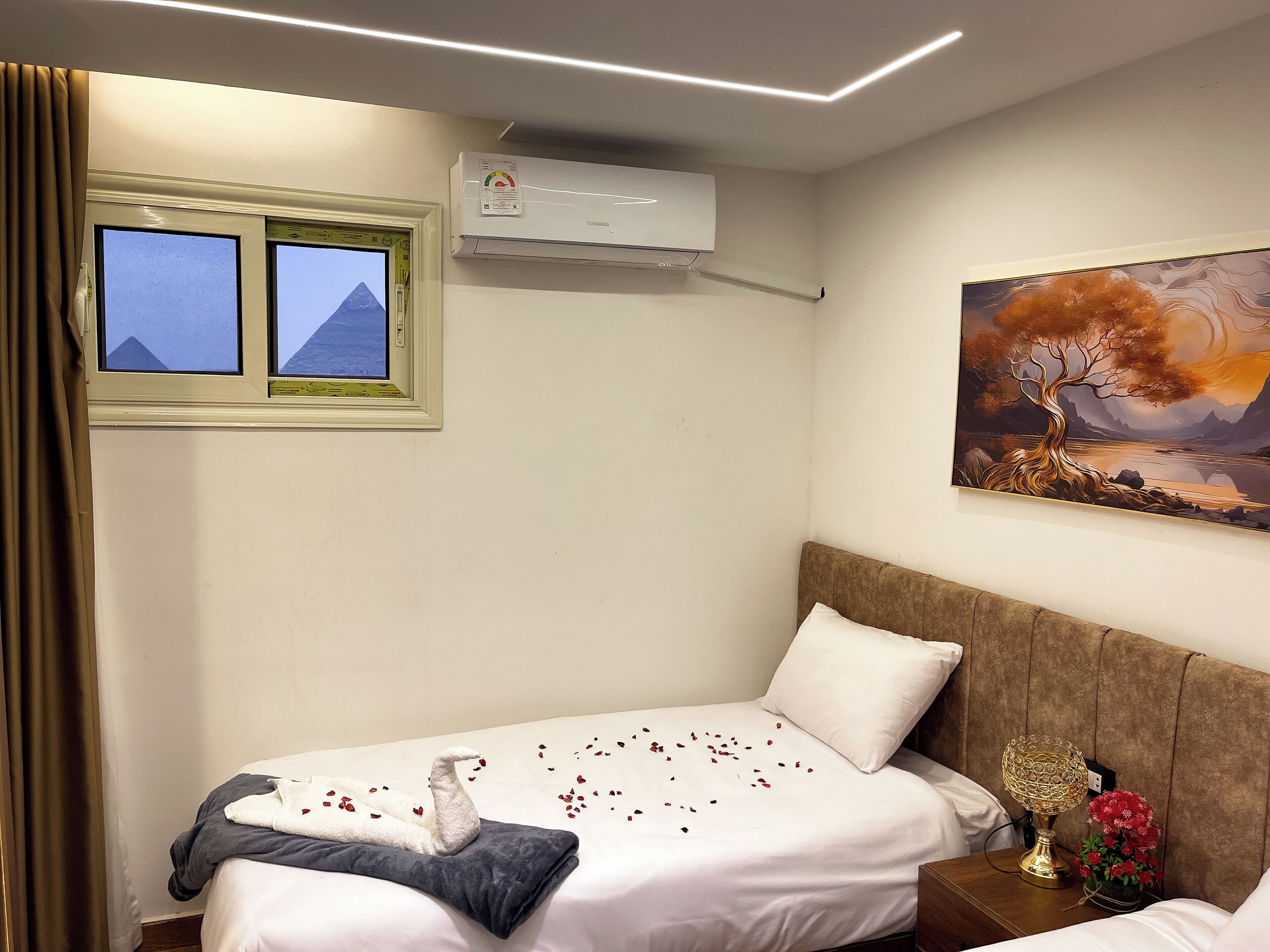 A hotel room with a bed, a nightstand, a wall-mounted air conditioner, a window with a view of pyramids, and a landscape painting.