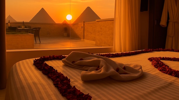 A hotel room with a view of the pyramids at sunset, a bed with rose petals, and a robe hanging on the wall.