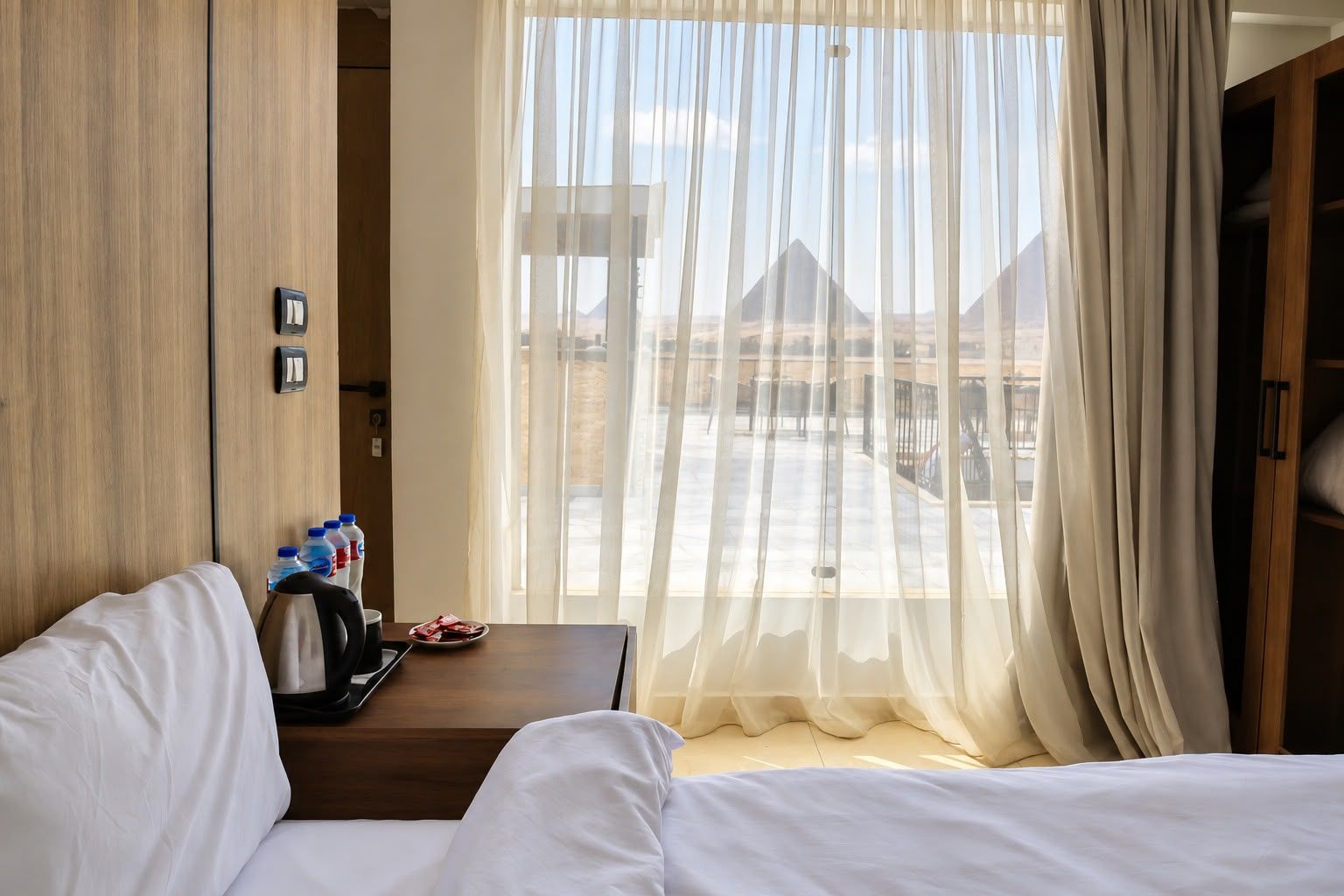 A hotel room with a bed, a small table with a coffee maker, a water bottle, and a plate of food. There is a window with sheer curtains and a view of pyramids.