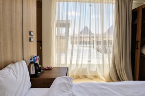 Habitación de hotel con cama, una mesita con cafetera, una botella de agua y un plato de comida. Se ve una ventana con cortinas translúcidas y vistas a pirámides.