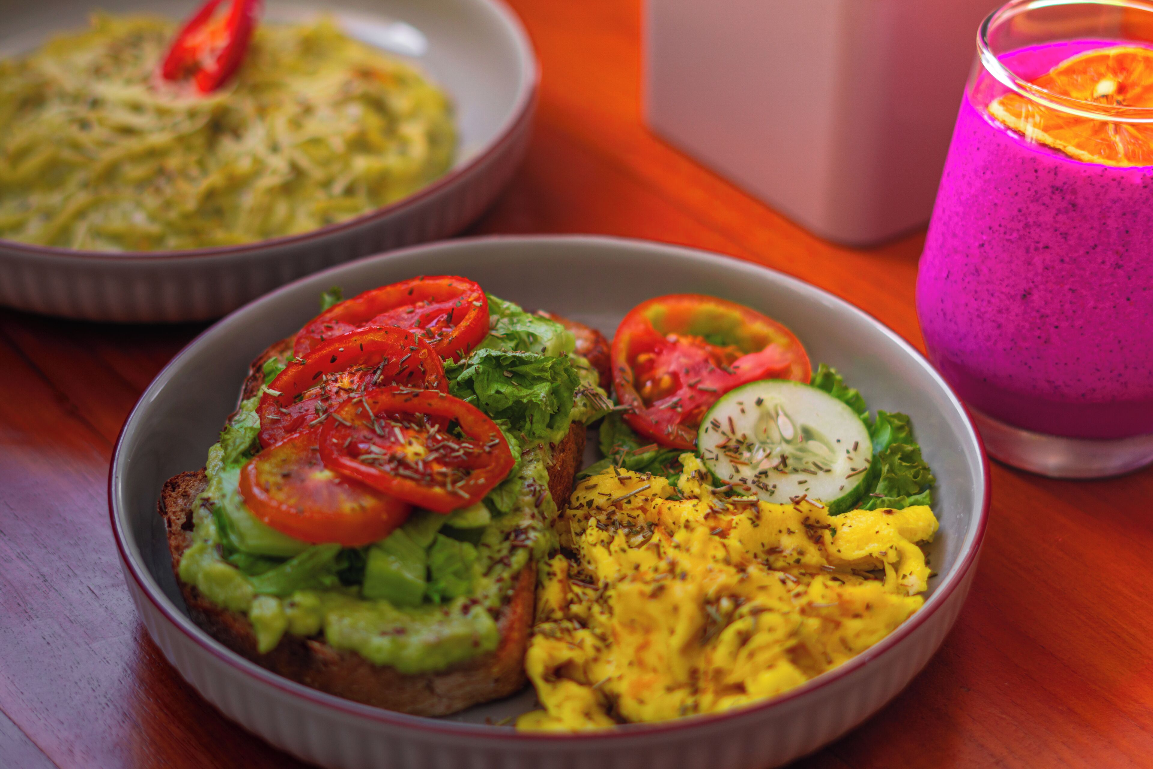 Uma refeição com torradas de abacate, ovos mexidos e um smoothie colorido.