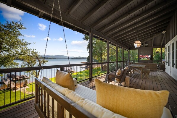 A covered deck with a swing chair, a dining table, and a view of a lake and mountains.