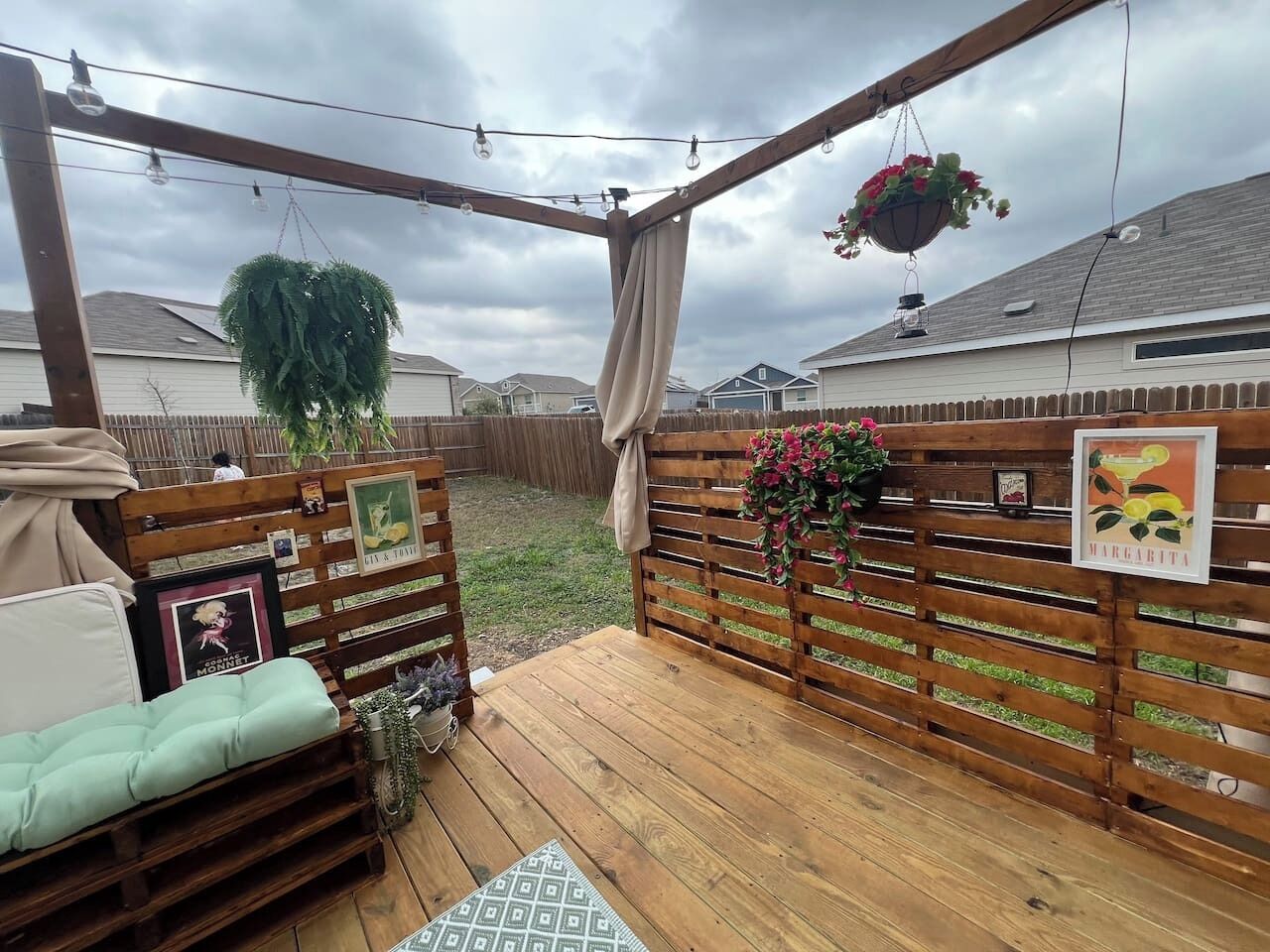 An outdoor deck with a wooden bench, a hanging planter, and a fence with a picture frame.