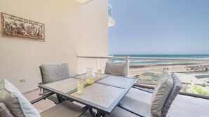 A balcony with a table and chairs, a painting on the wall, and a view of the beach and ocean.