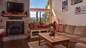 Een gezellige woonkamer met een stenen open haard, een groot raam met uitzicht op de natuur en een aan de muur bevestigde televisie.