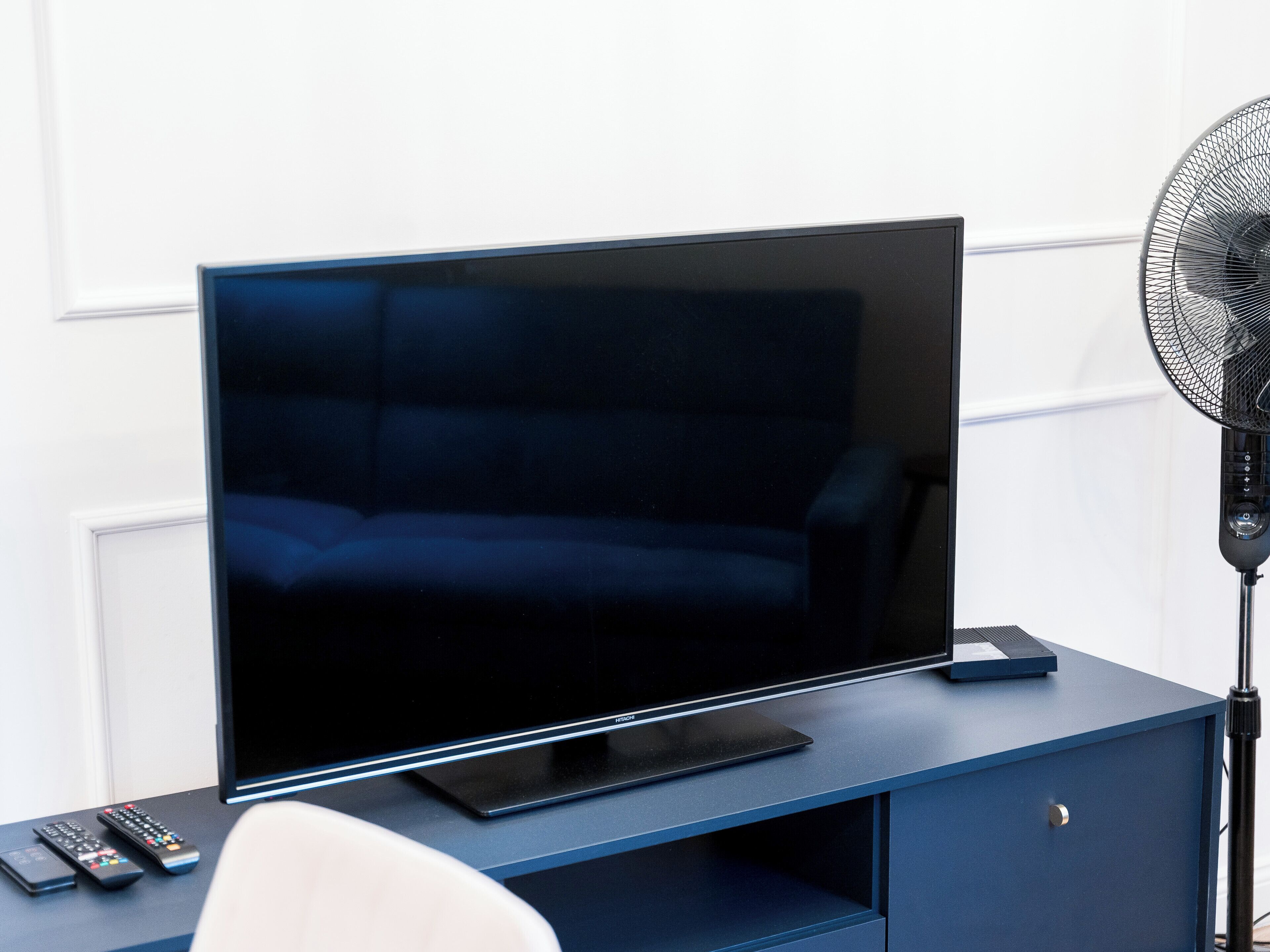 A modern living room with a flat-screen TV on a dark blue cabinet, a standing fan, and a white chair.