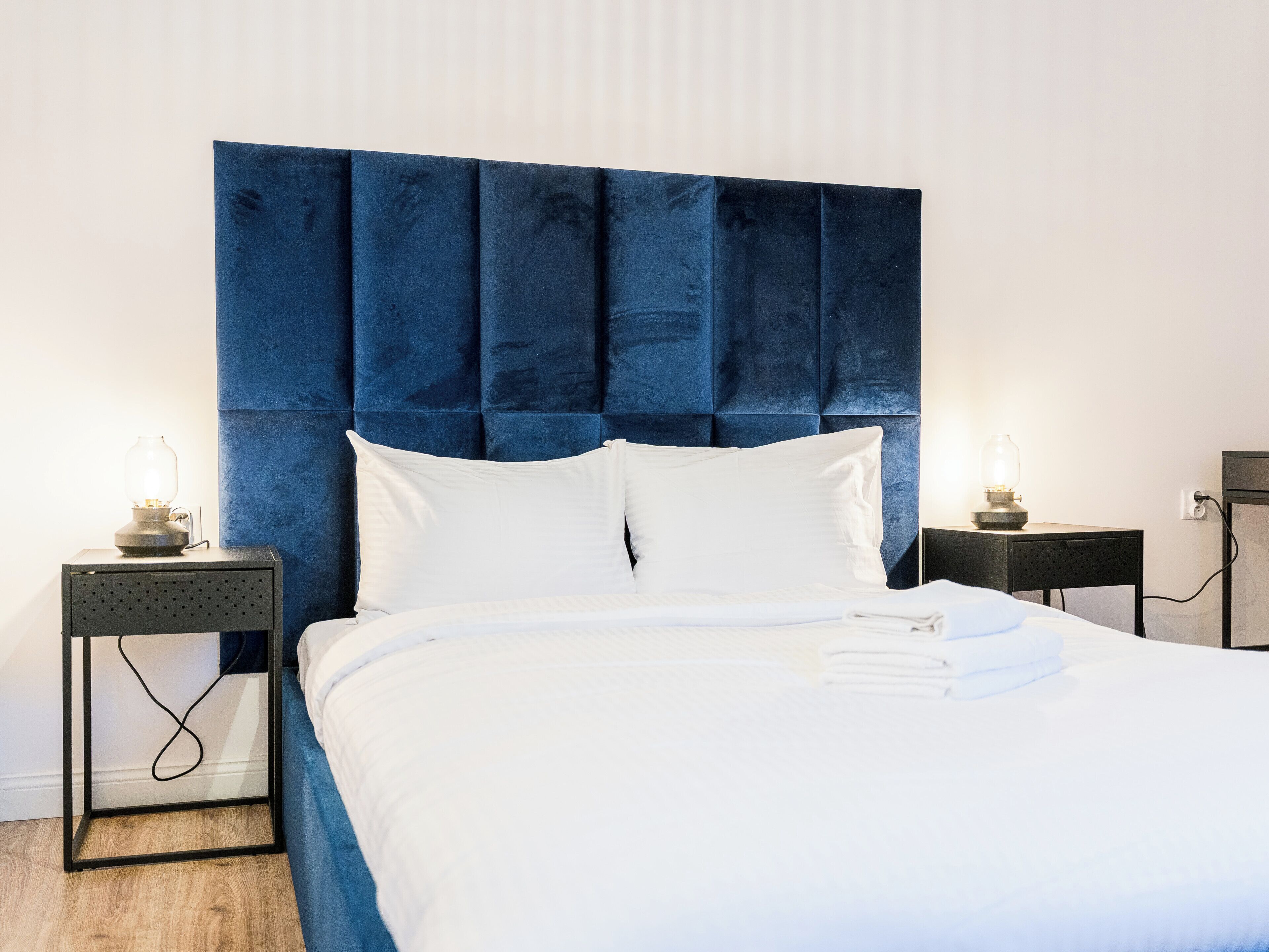 Una cama bien hecha con sábanas blancas, un cabecero tapizado en azul y dos mesitas de noche con lámparas.