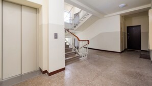 A clean, modern hallway with a polished stone floor, a wooden handrail on a staircase, and a door at the end. - Grójecka City Gateway Apartment (Warszawa)