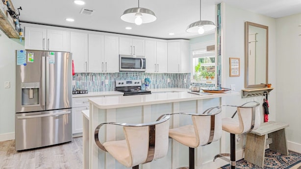 Una cocina moderna con armarios blancos, electrodomésticos de acero inoxidable, una isla central con taburetes altos e iluminación colgante.