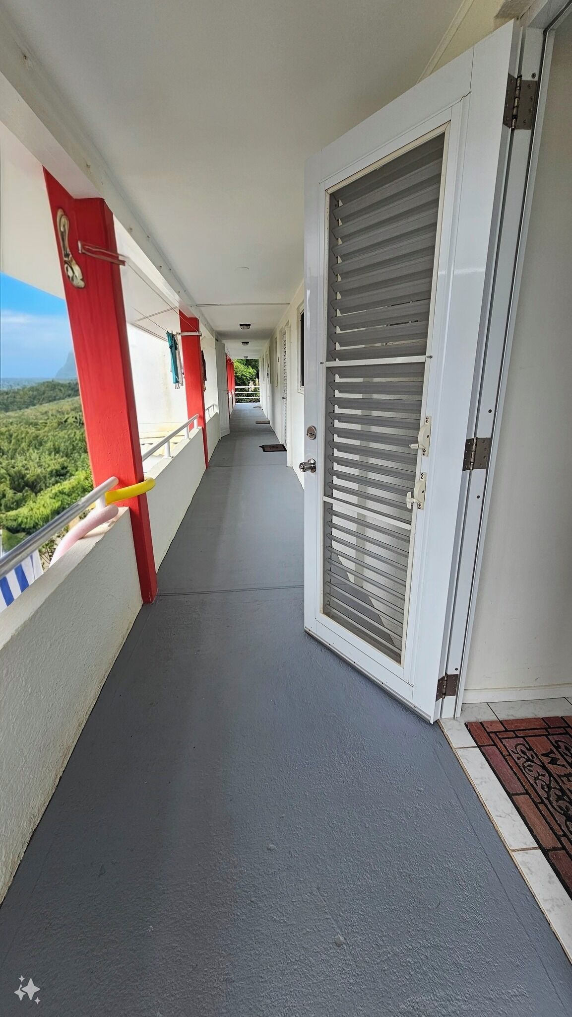 A corridor with a red pillar, a closed door with horizontal blinds, and a view of greenery outside.