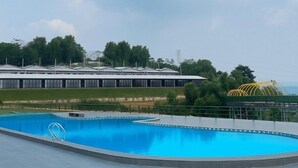 A swimming pool with a clear blue water, surrounded by a paved area and a building with a white roof in the background. - Wind Hill Mountain View Resort (Megamendung)