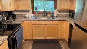 A kitchen with wooden cabinets, a granite countertop, a stainless steel sink, a stove, a microwave, and a window with a view of trees. - Charming Cabin, Lake Access! Backyard Bubbling Brook. Pets Welcome! (Denmark)