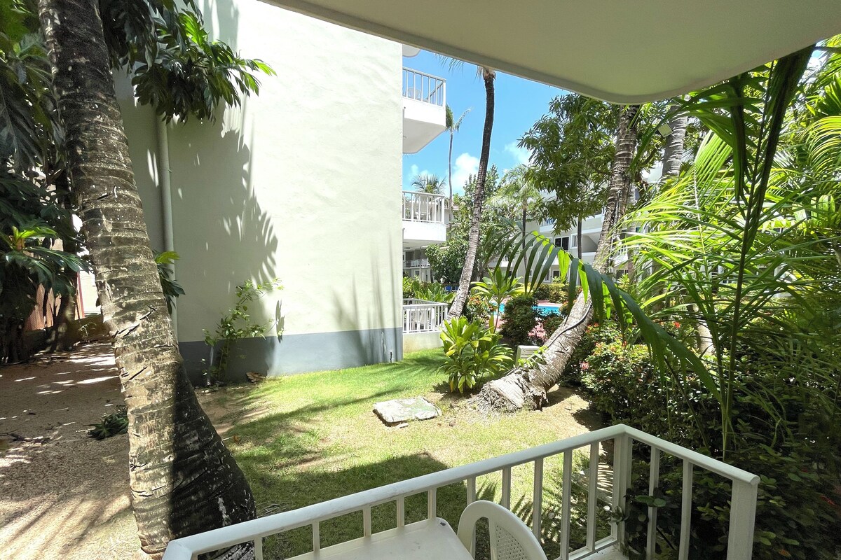 Un balcone con un tavolo e una sedia bianca, immerso nel verde rigoglioso e affacciato su un edificio dalla facciata bianca.