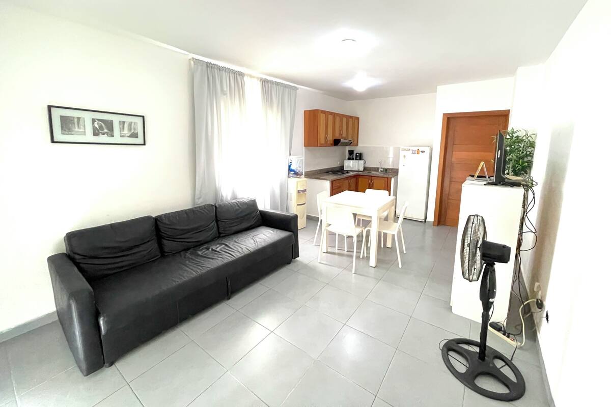 Een moderne woonkamer met een zwarte leren bank, een eettafel met stoelen, een keuken met houten kastjes, een witte koelkast en een staande ventilator.