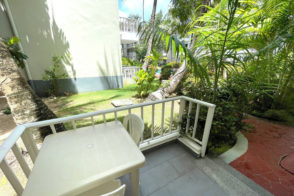 Un piccolo patio esterno con tavolo e sedie, immerso nel verde e con vista su un edificio sullo sfondo.