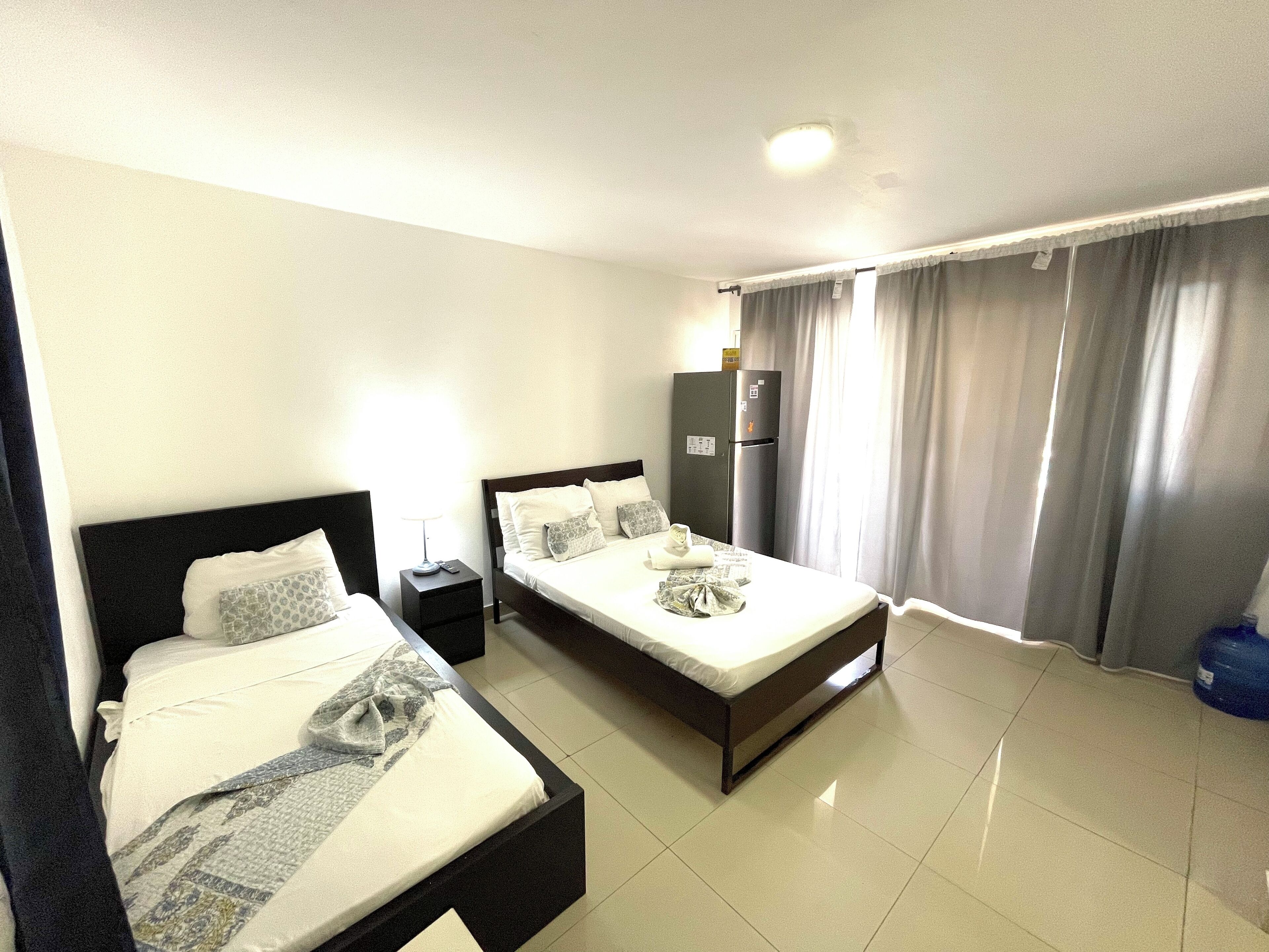 A hotel room with two beds, a refrigerator, a nightstand, and a window with curtains.